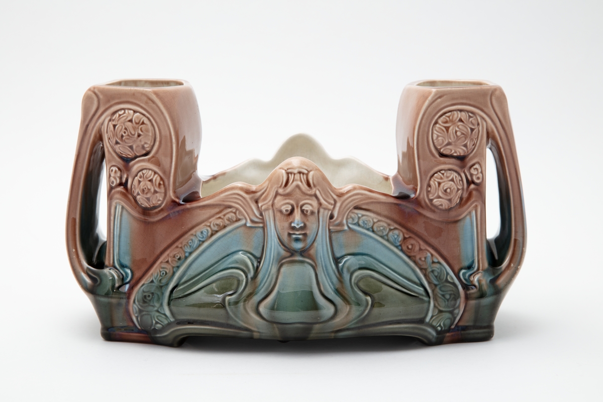 Avlang blomstervase (eller jardiniere) fra Egersunds Fayancefabriks Co. Merket E under bunnen. Jugendstil (designet av Andreas Ollestad 1898). Dekorert med plastiske masker, rosetter og abstrakt ornamentikk. Flytende glasur i sjøgrønt og blekt rosa farger utenpå, kremhvit inni. Gjennombrudt hank på hver kortside. Merket E under bunnen. Krakeleringer og noen få skader i glasuren.