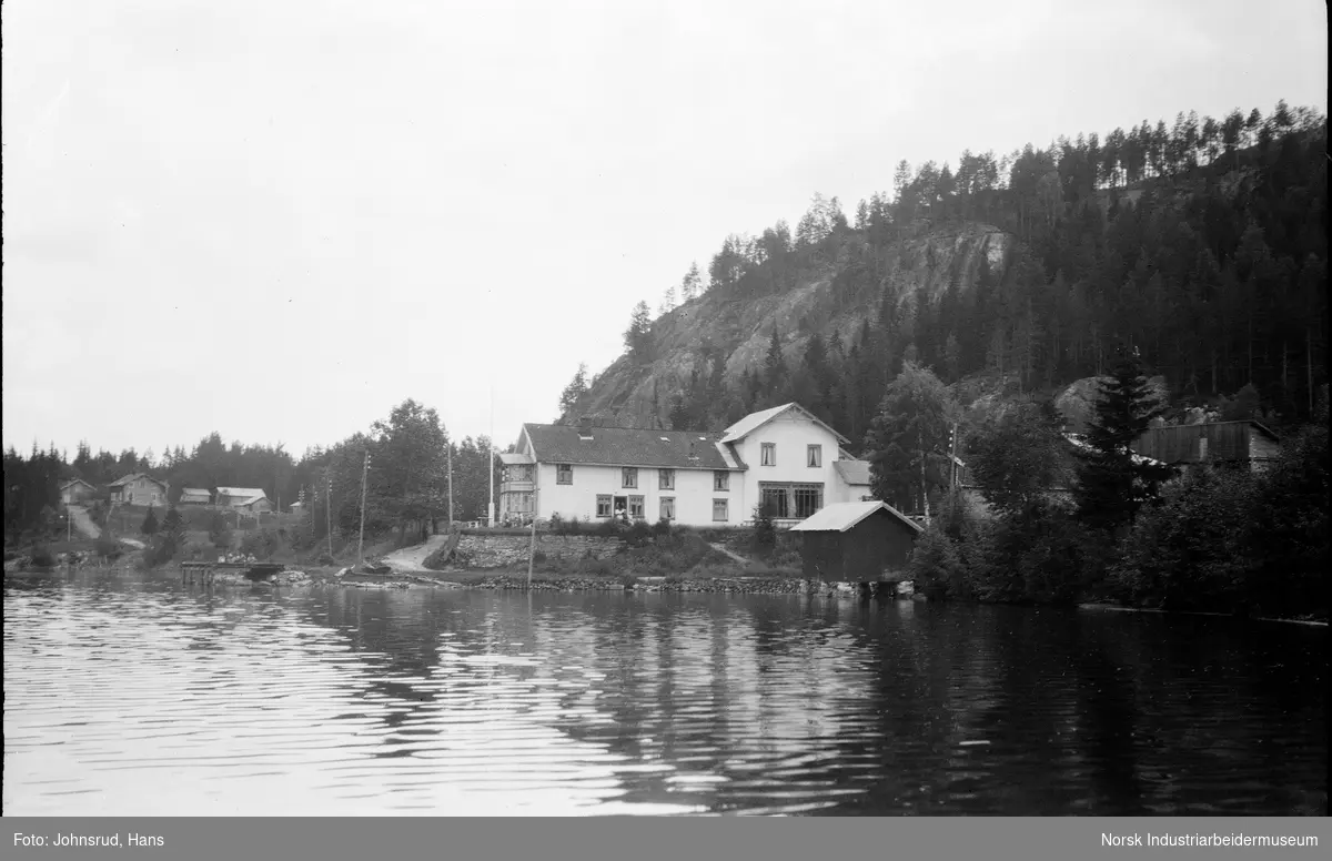 Hotell på Tinnoset sett fra vannet. Mennesker er synlige foran hotellet. Foran hotellet er det et lite båthus, og til venstre er den en liten brygge, I bakgrunnen er det en liten ås på høyresiden og flere bygninger på venstresiden.