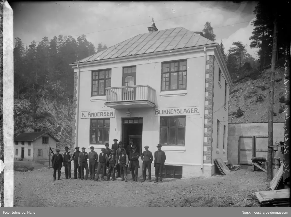 Bilde av en gruppe foran butikken til blikkenslager Andersen. Det står femten menn i inngangspartiet til bygningen. Bygningen er et to etasjers murbygg med påskriften "K. Andersen" og "Blikkenslager." over førsteetasje vinduene. Over inngangspariet er det en balkong. I venstre side av bildet er det en bygning.