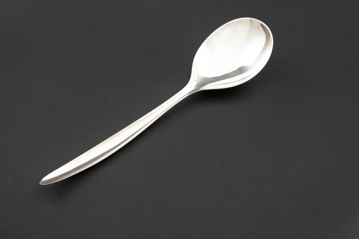 Kompottskje i sølvplett. Stanset ut i jevntykk plate. Ovalt skjeblad med fint kurvet overgang til skaft. Bladets fordypning glir jevnt over i fordypning langs skaftets innside og gir skjeen en utpreget konveks og konkav side. Glatt overflate uten dekor.