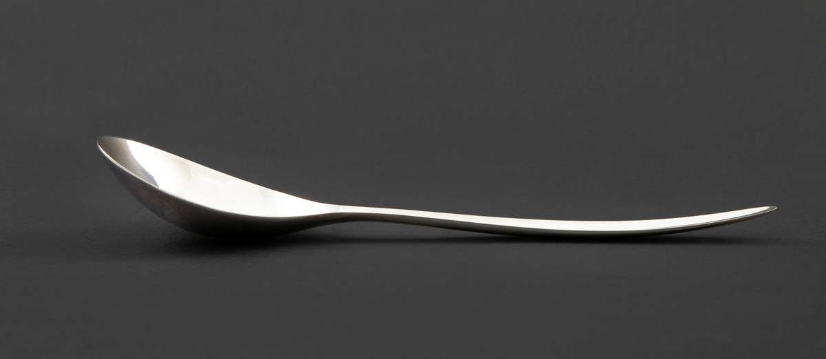 Kompottskje i sølvplett. Stanset ut i jevntykk plate. Ovalt skjeblad med fint kurvet overgang til skaft. Bladets fordypning glir jevnt over i fordypning langs skaftets innside og gir skjeen en utpreget konveks og konkav side. Glatt overflate uten dekor.