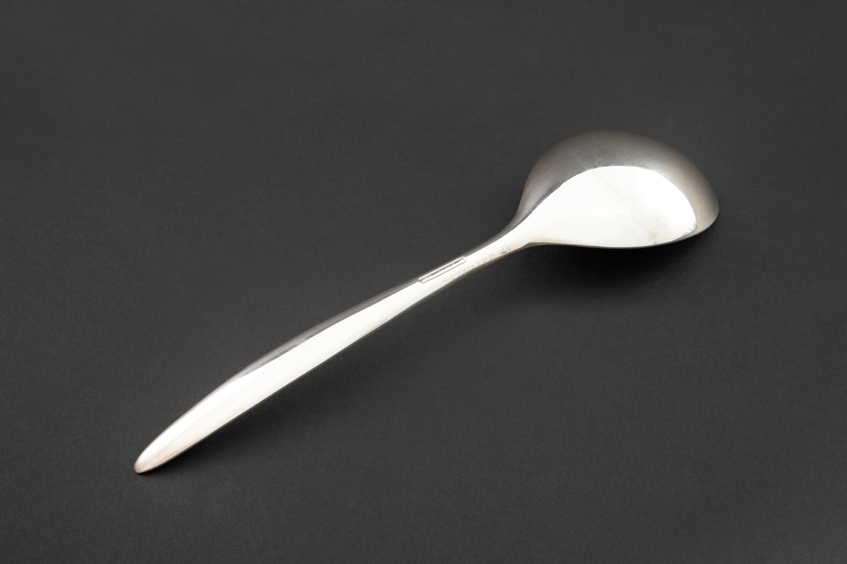 Kompottskje i sølvplett. Stanset ut i jevntykk plate. Ovalt skjeblad med fint kurvet overgang til skaft. Bladets fordypning glir jevnt over i fordypning langs skaftets innside og gir skjeen en utpreget konveks og konkav side. Glatt overflate uten dekor.