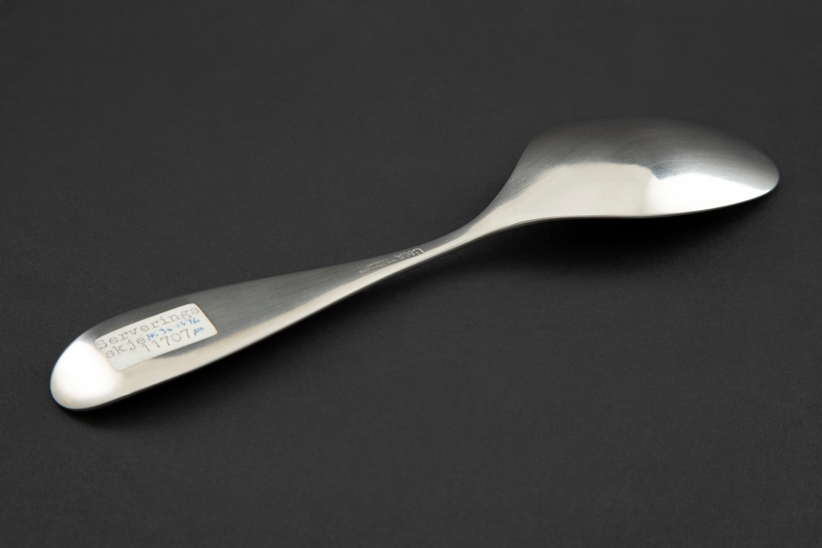 Serveringsskje i mattpolert rustfritt stål. Ovalt skjeblad med smal legg og dråpeformet skaft med oppbøyd, avrundet ende.