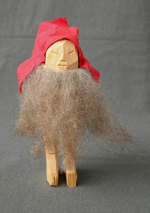 Jultomte av trä med täljd kropp, ögon, näsa och mun, grått fårskinn till skägg och ett rött huckle av pappersservett fastspikat på huvudet med nubb.