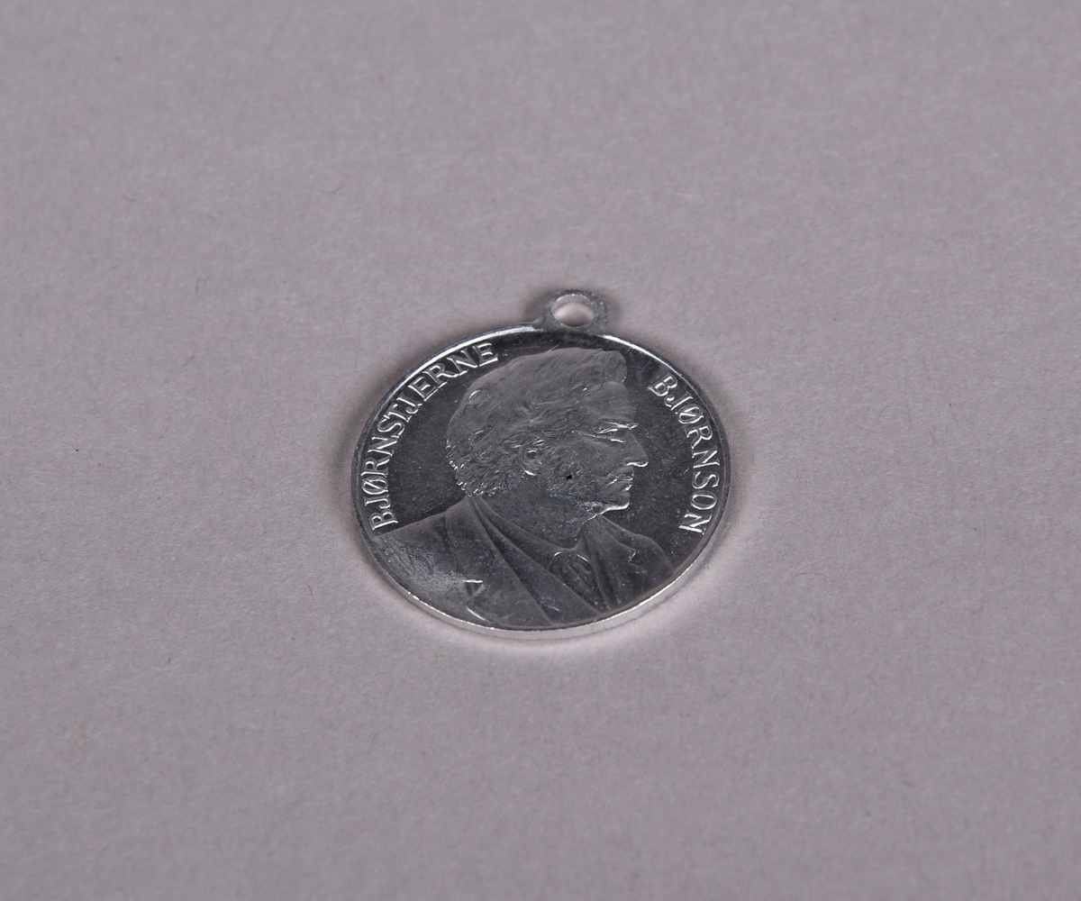 Rund medalje med hempe øverst. Motiv av Bjørnstjerne Bjørnson på den ene siden, og på den andre siden en dame i kjole, i hånden holder hun et skjold med det norske riksvåpnet.
