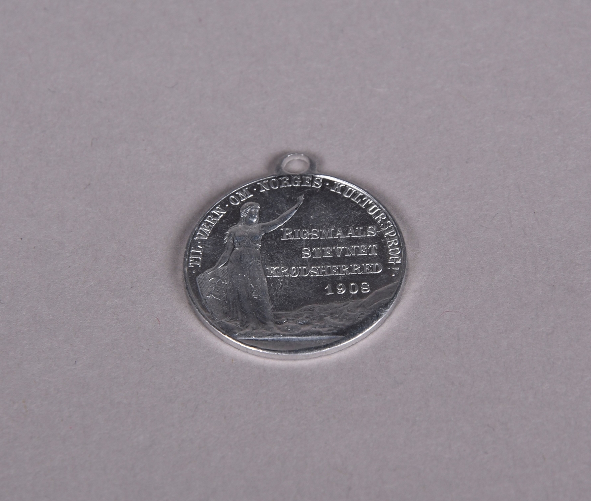 Rund medalje med hempe øverst. Motiv av Bjørnstjerne Bjørnson på den ene siden, og på den andre siden en dame i kjole, i hånden holder hun et skjold med det norske riksvåpnet.
