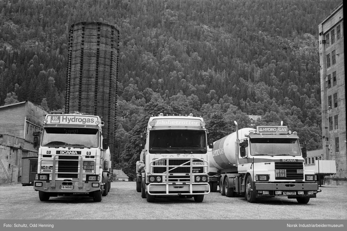 Lastebiler til transport av gass står parkert på fabrikkområdet. Det står Hydrogas på kjøretøyene. Bak er det et syretårn.