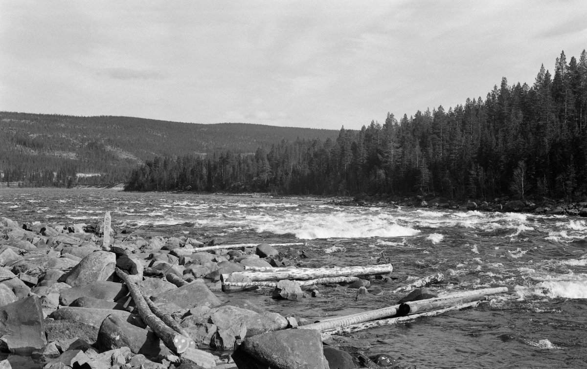 Glomma eller Glåma, fotografert en vårdag i 1964, et sted i Alvdal i Nord-Østerdalen. Elva gikk flomstor, til dels gjennom kvitskummende stryk, gjennom et ubebodd område med mye barskog. Til venstre i forgrunnen ser vi en steinete elvebredd der noen tømmerstokker (skipvirke) hadde strandet. På dette stedet hadde noen reist en bordstubb med innskriften «G.B.4» på et bless i den øvre enden. Bildet ble antakelig tatt for å dokumentere områder der det var hindringer for tømmerfløtinga i Glomma. Dokumentasjonen ble gjennomført med tanke på såkalte «elveforbedringer», fysiske tiltak som skulle endre elvetopografien til beste for fløtinga i  vassdraget. «G.B.4» refererer antakelig til ei liste med foreslag om hva som burde gjøres på ulike lokaliteter.