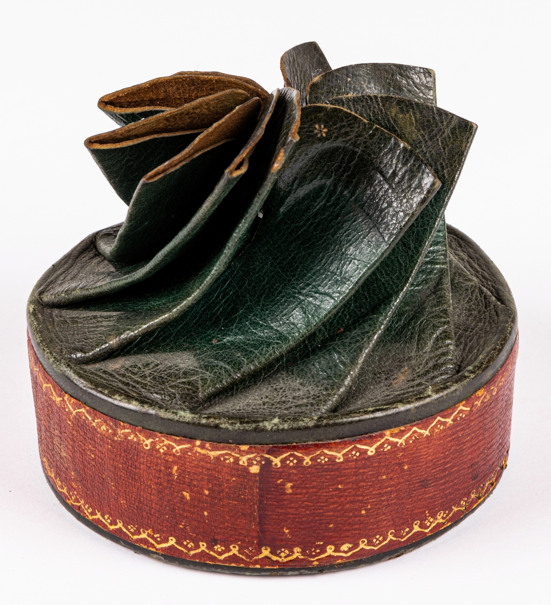 Tobaksask, låg, rund pappask, med botten av papp och lock av skinn skuret med snibbar och vridet koncentriskt så att snibbarna bilda en tiouddig figur. Sidorna röda, ovansidan skinnet grönt. Smala guldbårder på askens sidor och texten "Joh. Olof Norin", samt små guldstjärnor på ovansidan skinnet. Skinnet är saffian.
