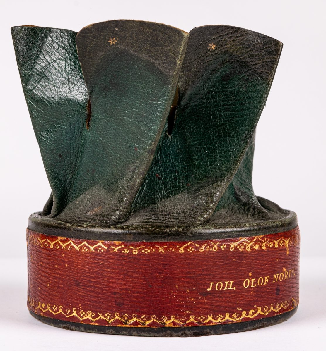 Tobaksask, låg, rund pappask, med botten av papp och lock av skinn skuret med snibbar och vridet koncentriskt så att snibbarna bilda en tiouddig figur. Sidorna röda, ovansidan skinnet grönt. Smala guldbårder på askens sidor och texten "Joh. Olof Norin", samt små guldstjärnor på ovansidan skinnet. Skinnet är saffian.