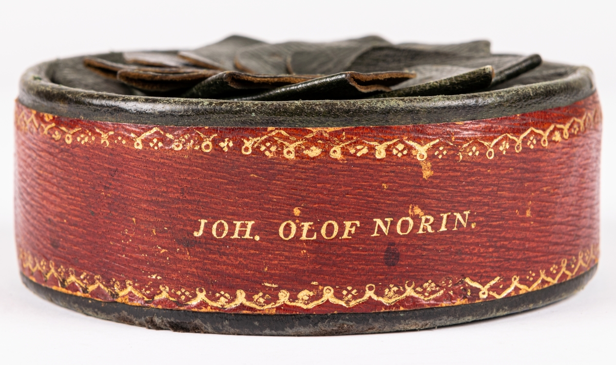 Tobaksask, låg, rund pappask, med botten av papp och lock av skinn skuret med snibbar och vridet koncentriskt så att snibbarna bilda en tiouddig figur. Sidorna röda, ovansidan skinnet grönt. Smala guldbårder på askens sidor och texten "Joh. Olof Norin", samt små guldstjärnor på ovansidan skinnet. Skinnet är saffian.