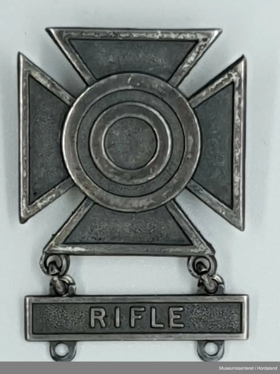 Medalje eller ferdighetsmerke til å feste på uniform. Hoveddelen er et likebeinet kors, nærmest fire trekanter som stikker ut fra sentrum. Medaljens sentrum viser relieff av fire ringer utenfor hverandre, som en blink. Overflaten veksler mellom glatt og ruglete, som også gir effekt i det visuelle utrykket. Festet under korset med med løse ringer, henger en plate med inkripsjonen RIFLE, under platen er det to ringer, som muliggjør feste av flere plater som viser til ulike ferdigheter og våpentyper. 