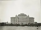 Växjö högre allmänna läroverk, ca 1905. Ett antal pojkar är ute och marscherar m.m. på skolgården, betittade av en flicka. 
Till vänster skymtar borgmästare Bergendahls hus i hörnet Trädgårdsgatan/Parkgatan och bakom det Flickskolan. 
Till höger skymtar man bokhandlare Quidings hus på Södra Järnvägsgatan.