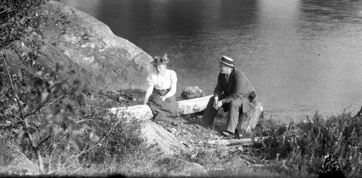 Kvinne og mann ved et lite gårdsbruk ved sjøen. Bratt hei bak. Båthus og pram. - kanskje ved Lindvika?