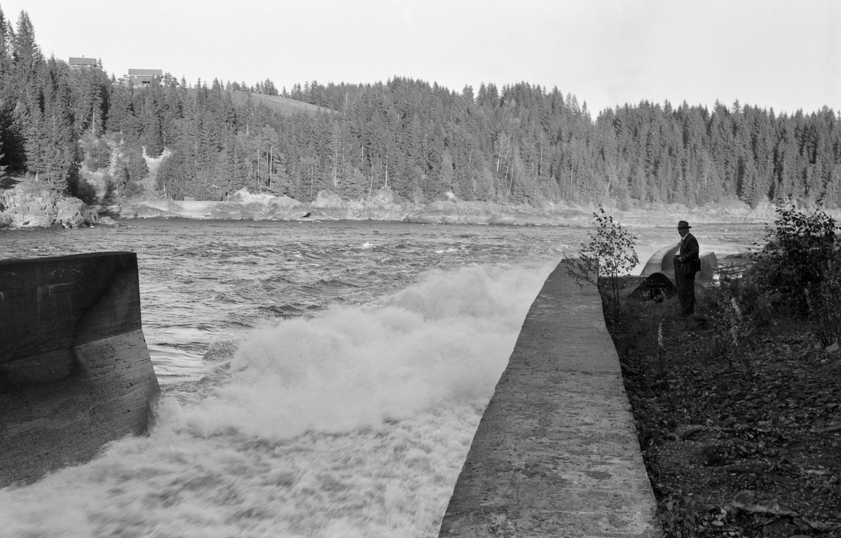 Fra utløpet av tømmerkanalen som i andre halvdel av 1920-åra ble bygd på sørsida av dammen og kraftverket ved Vamma i Nedre Glomma. Den øvre delen av kanalen ble sprengt ned i fjellet, der botnen og sideflatene ble forblendet med tømmer eller betong for å sikre at fløtingsvirket ikke skulle sette seg fast. Den nedre delen av kanalen ble støpt i betong, noe som jo innebar at løpet ble forholdsvis glatt. Her var hellinga forholdsvis bratt, noe som framgår av dette bildet ved at vannet var kvitskummende og opprørt der hvor kanalen møtte elva nedenfor dammen og kraftstasjonen. Dette fotografiet viser også hvordan det var støpt en betongskjerm, en skådam, som skulle forebygge at tømmeret som kom ut av renna ble presset inn mot land. En dresskledd mann med hatt betraktet strømkavet ved rennas utløp da dette bildet ble tatt. På bakkekammen på motsatt side av elveløpet skimter vi et par av bygningene på gardsbruket Fosser på Askim-sida av elva.