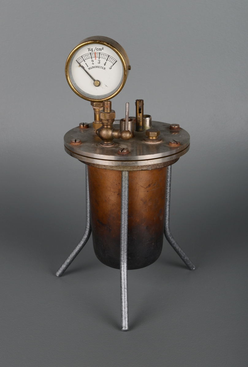 En trykktank av jern bestående av en kjeleformet tank som står i et rundt stativ med tre tynne bein. På toppen er det en måler av glass og messing for manometeret, som brukes inne i tanken til å måle trykk (kg/cm2). Måleskalaen går fra 0 til 5. Tanken er sylinderformet og lokket er skrudd fast med seks store skruer.