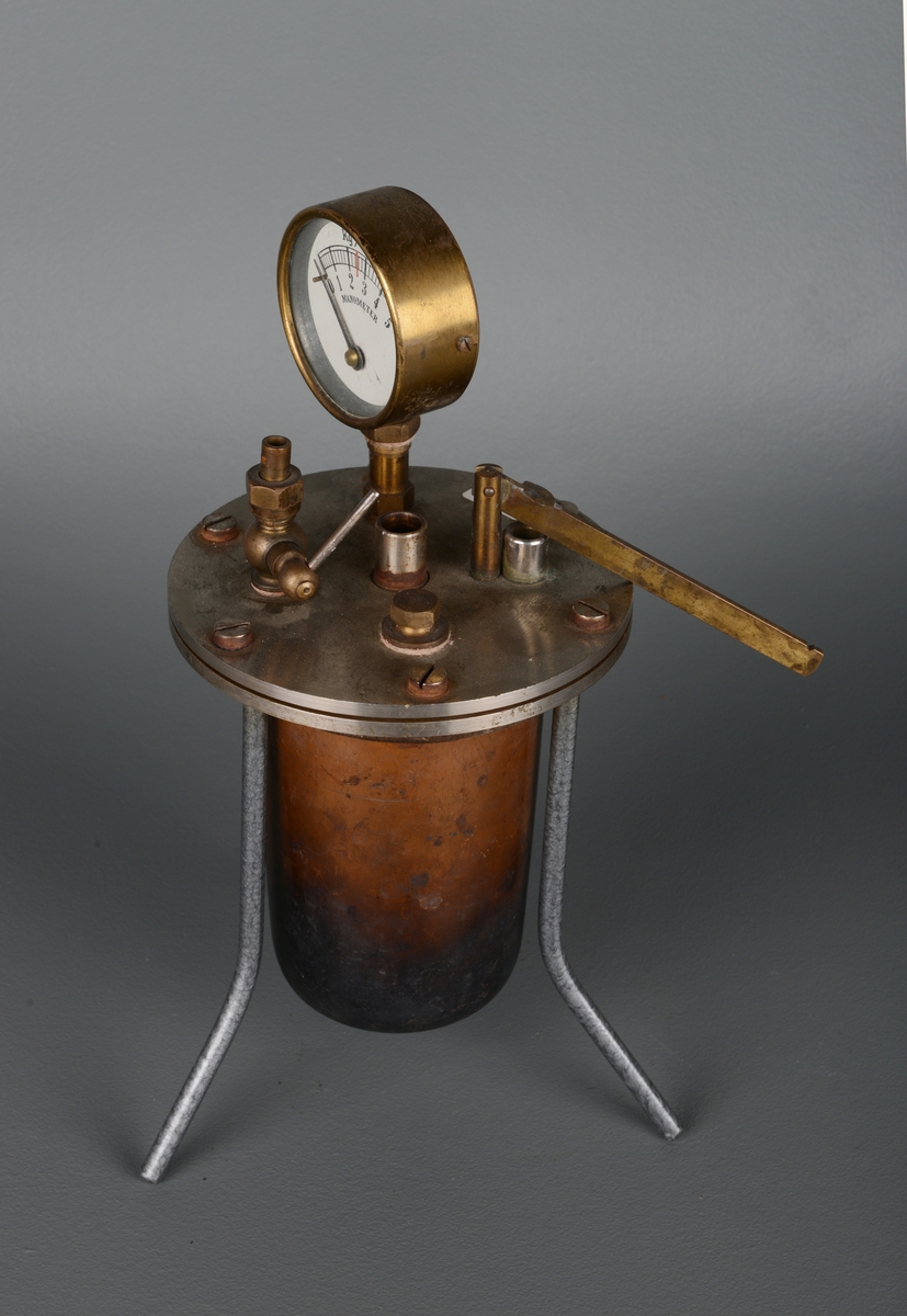 En trykktank av jern bestående av en kjeleformet tank som står i et rundt stativ med tre tynne bein. På toppen er det en måler av glass og messing for manometeret, som brukes inne i tanken til å måle trykk (kg/cm2). Måleskalaen går fra 0 til 5. Tanken er sylinderformet og lokket er skrudd fast med seks store skruer.