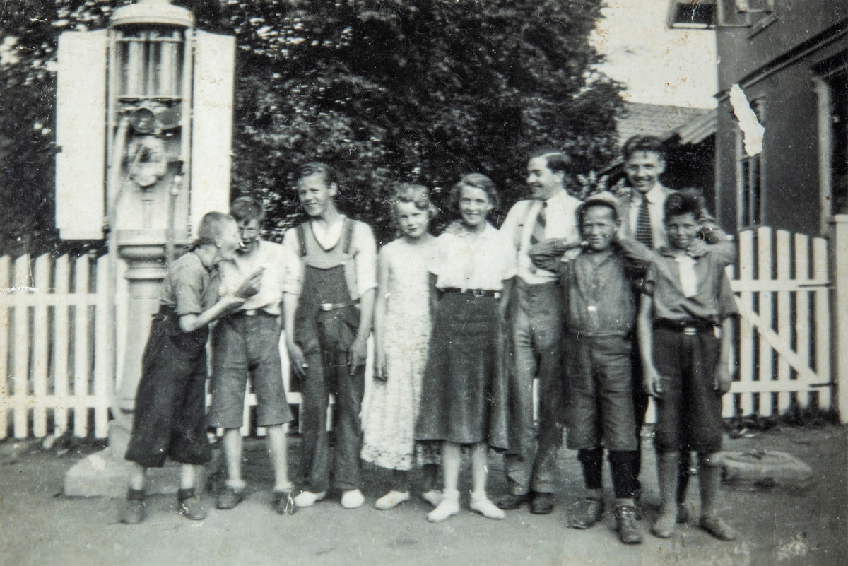 Gruppe 9 ungdommer, ved siden av bensinpumpen utenfor kolonialforretningen til Kasper Engh i Bekkelaget en gang på 1930-tallet. Ottestad, Stange. De to mennene bak til høyre, var handelsbetjenter i butikken til Kasper Engh. Handelsbetjent Bjarne Finstad, som senere åpnet egen forretning i Stange. Og handelsbetjent ? Martinsen, som senere åpnet egen butikk på Nes, Ringsaker. De øvrige personene er ukjente.