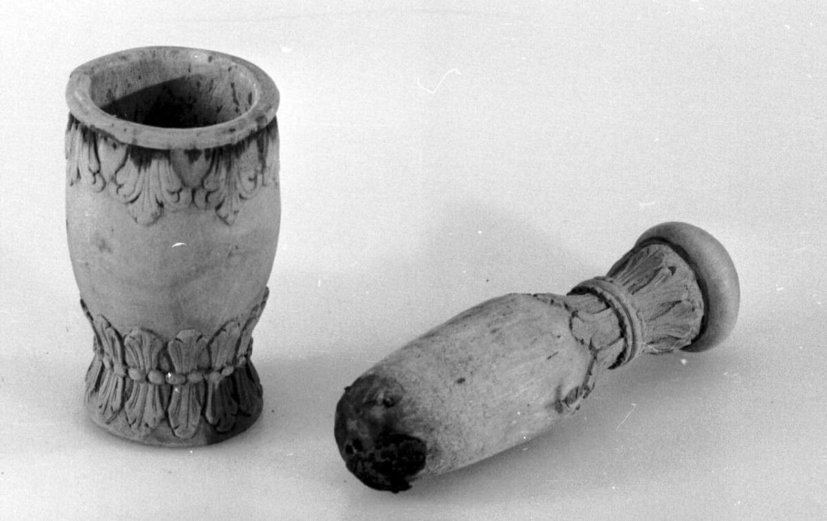 Morter og pistill av tre. Utskjæringer av palmetter både øverst og nederst på morter og øverst på pistill. Palmettene er bundet av perlerad nederst. Det er et hull nederst i morteren. Nederst på pistillen har det vært festet et seks-bladet metallbeslag, nå er det kun to blader tilbake. Det sitter igjen fire spiker fra beslagene som er borte.