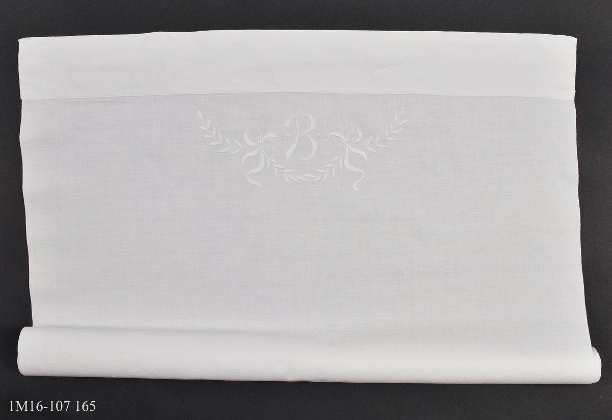 Lakan, vitt, dekorerat med monogram, B med en bladsling med två rosetter under. Broderat med plattsöm. Sytt av mormor Beda Skaar till mor Brita.