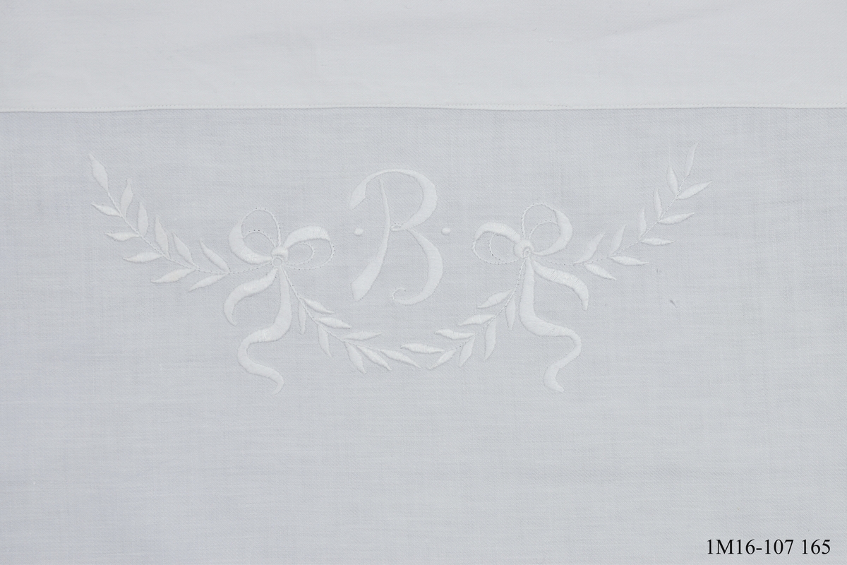 Lakan, vitt, dekorerat med monogram, B med en bladsling med två rosetter under. Broderat med plattsöm. Sytt av mormor Beda Skaar till mor Brita.