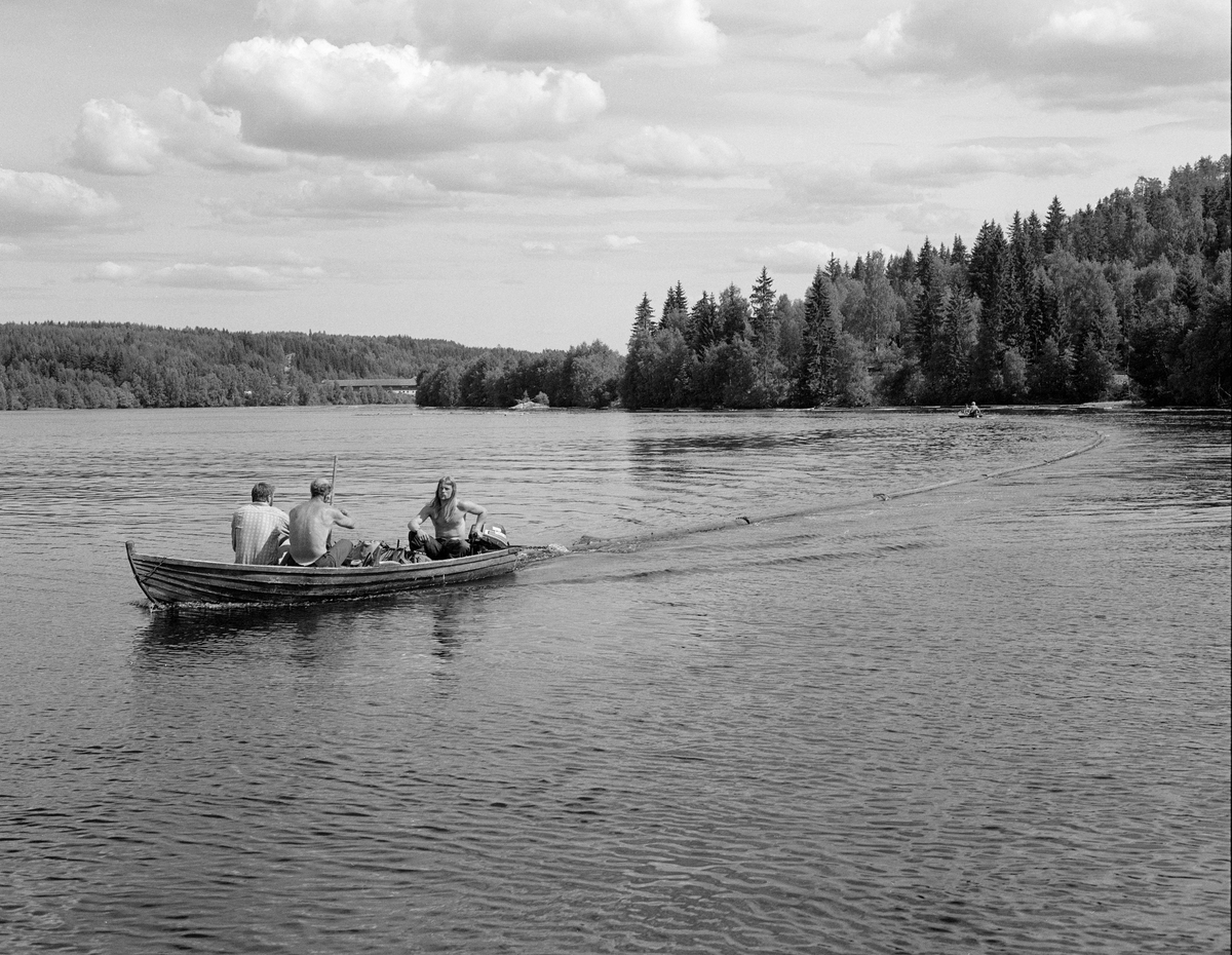 Sluttrensk i Glomma i juni 1985. Fotografiet viser to klinkbygde trebåter med påhengsmotorer på et relativt stilleflytene elveløp. Bak den fremste båten hang det ei oppsamlingslense som ble brukt til å trekke med seg gjenliggende løstømmer. Kenth Mokkelbost, som ledet arbeidet med sluttrensken, førte båten. 1985 var det siste året det foregikk ordinær fløting i Glommavassdraget. Fotografiet er tatt mellom Hanstadfossen og Skjefstadfossen i Elverum kommune. Vi skimter driftsbygningen på garden Østerhaug i bakgrunnen.