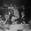 Christian Diors besök på NK, Nordiska kompaniet i Stockholm, februari 1957. Han bjuds på en visning av plagg uppsydda av tyger från NK:s textilkammare av Kurt Jacobsson och Pelle Lundgren. Mannekäng visar plagg. Vid bordet sitter Dior tillsammans med Astrid Sampe.