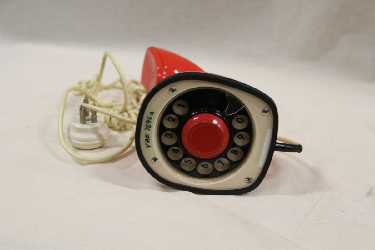 Rød teleofn med tall-dreieskive og summetonebryter under på foten. Ledning går ut fra baksiden på bunnen. Formen på telefonen kan minne om hodet på en Kobraslange.