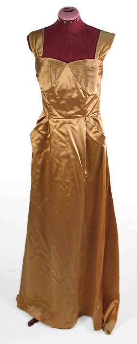 Klänning av guldfärgat atlassiden.
Liv: Kupformat liv med lagda veck mot överkanten. 4 st små släta våder, kilformat mellan kuporna. 2 st breda axelband med 1 veck i framkanten. Bystdelen 2 st lagda veck. Insydd bysthållare. Bakstycket i 4 st släta delar med insyningar. Hänghällor under ärmarna. Livet fodrat med vitt tätvävt bomullstyg. 6 st insydda spiraler.
Mått i cm: Axellängd 37, bredd 7.
Kjol: 2 st raka våder framtill. Slät mitt fram med ett stort fickliknande veck samt ett litet veck på var sida framstycket. Bakvåden snedklippt med mycket vidd samt släp. 36 cm långt blixtlås baktill. 
Mått: Kjolvidd 452 cm.