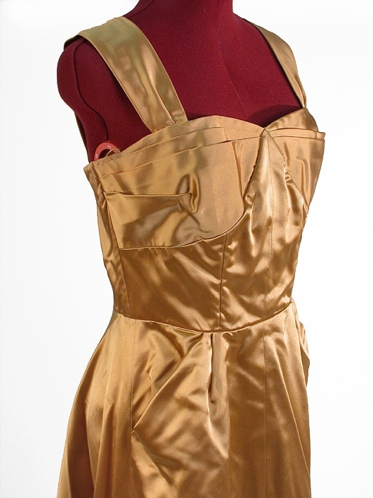 Klänning av guldfärgat atlassiden.
Liv: Kupformat liv med lagda veck mot överkanten. 4 st små släta våder, kilformat mellan kuporna. 2 st breda axelband med 1 veck i framkanten. Bystdelen 2 st lagda veck. Insydd bysthållare. Bakstycket i 4 st släta delar med insyningar. Hänghällor under ärmarna. Livet fodrat med vitt tätvävt bomullstyg. 6 st insydda spiraler.
Mått i cm: Axellängd 37, bredd 7.
Kjol: 2 st raka våder framtill. Slät mitt fram med ett stort fickliknande veck samt ett litet veck på var sida framstycket. Bakvåden snedklippt med mycket vidd samt släp. 36 cm långt blixtlås baktill. 
Mått: Kjolvidd 452 cm.