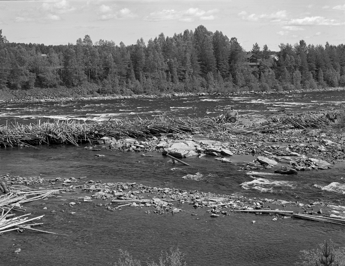 En stor tømmerhaug som hadde bygd seg opp i elveløpet nedenfor Skjefstadfossen i Heradsbygda i Elverum løsnes ved hjelp av en hjullaster. Fotografiet ble tatt sommeren 1984, på et tidspunkt da det var lav vannstand nedenfor kraftverksdammen, Arbeidet ble gjort som et ledd i «sluttrensken», som innebar at tømmerstokker som hadde strandet langs elvebreddene eller ved steinskjær og på grusører ute i elveløpet skulle bringes tilbake i det strømmende vannet igjen. Slikt arbeid ble vanligvis utført manuelt, av menn med fløterhaker.