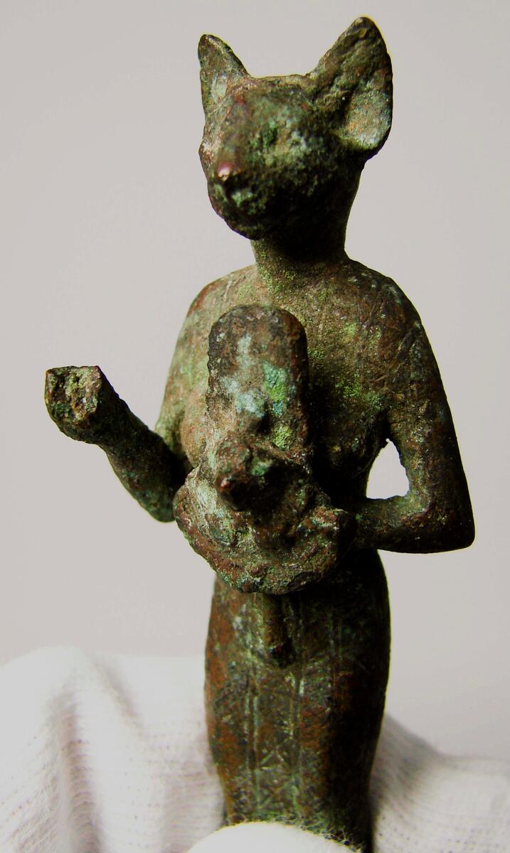 Figurin av brons, Egypten- Egyptisk gudabild föreställande Gudinnan Bastet, symboliserad som en katt. Daterad till Setiden, 664-525 f.kr. Längd: 9 cm, Höjd: 2 cm, Bredd: 2,7 cm.  Gudinnan Bastet har här avbildats med ett katthuvud på en kvinnokropp. I sin vänstra hand håller hon en ”aegis”, en skyddssköld med ett katt- eller Sekhmetansikte (krigets och solens gudinna med lejonhuvud på en kvinnorkropp) med en bred krage under. I sin högra hand har hon troligtvis hållit ett ”sistrum”, ett musikinstrument med små metallplattor vilka rasslade när man skakade instrumentet. Sistrumet är dock bortslaget. Statyetten har antagligen varit fastsatt på en träsockel.; ; Under Sentiden tillverkades små bronsfigurer i stora mängder och antagligen är denna statyett ett exempel på dessa. De skänktes därefter som votivgåvor till olika tempel. Tillverkningstid: 664-525 k Kr.