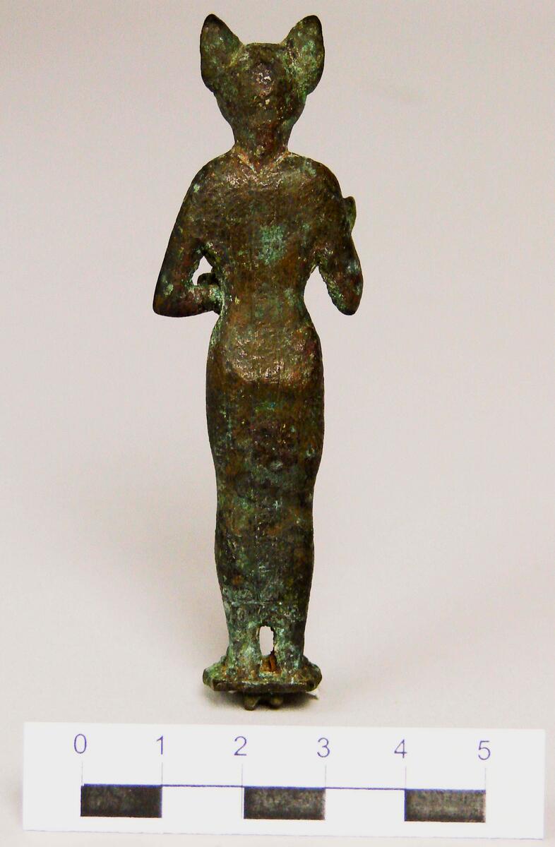 Figurin av brons, Egypten- Egyptisk gudabild föreställande Gudinnan Bastet, symboliserad som en katt. Daterad till Setiden, 664-525 f.kr. Längd: 9 cm, Höjd: 2 cm, Bredd: 2,7 cm.  Gudinnan Bastet har här avbildats med ett katthuvud på en kvinnokropp. I sin vänstra hand håller hon en ”aegis”, en skyddssköld med ett katt- eller Sekhmetansikte (krigets och solens gudinna med lejonhuvud på en kvinnorkropp) med en bred krage under. I sin högra hand har hon troligtvis hållit ett ”sistrum”, ett musikinstrument med små metallplattor vilka rasslade när man skakade instrumentet. Sistrumet är dock bortslaget. Statyetten har antagligen varit fastsatt på en träsockel.; ; Under Sentiden tillverkades små bronsfigurer i stora mängder och antagligen är denna statyett ett exempel på dessa. De skänktes därefter som votivgåvor till olika tempel. Tillverkningstid: 664-525 k Kr.
