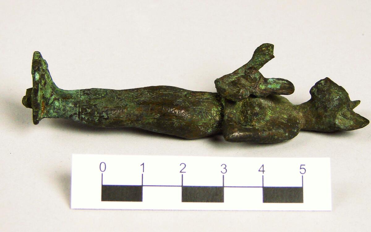 Figurin av brons, Egypten- Egyptisk gudabild föreställande Gudinnan Bastet, symboliserad som en katt. Daterad till Setiden, 664-525 f.kr. Längd: 9 cm, Höjd: 2 cm, Bredd: 2,7 cm.  Gudinnan Bastet har här avbildats med ett katthuvud på en kvinnokropp. I sin vänstra hand håller hon en ”aegis”, en skyddssköld med ett katt- eller Sekhmetansikte (krigets och solens gudinna med lejonhuvud på en kvinnorkropp) med en bred krage under. I sin högra hand har hon troligtvis hållit ett ”sistrum”, ett musikinstrument med små metallplattor vilka rasslade när man skakade instrumentet. Sistrumet är dock bortslaget. Statyetten har antagligen varit fastsatt på en träsockel.; ; Under Sentiden tillverkades små bronsfigurer i stora mängder och antagligen är denna statyett ett exempel på dessa. De skänktes därefter som votivgåvor till olika tempel. Tillverkningstid: 664-525 k Kr.