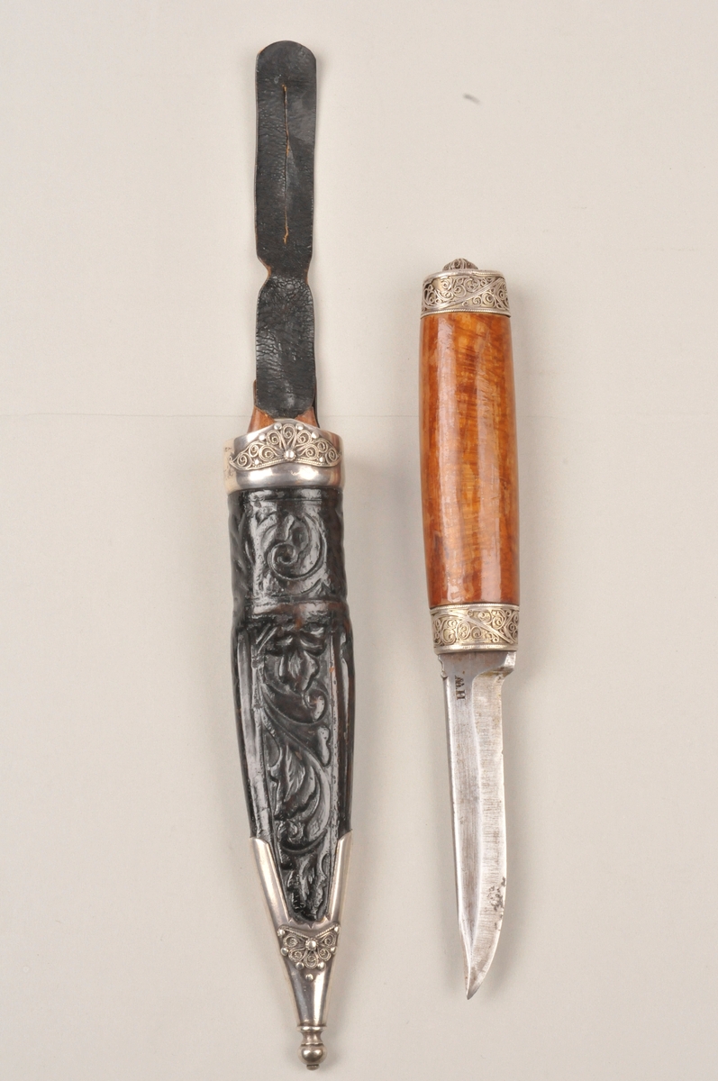 Slirekniv med sylvholkar oppe og nede på både skaft og slire, dekorera med filigransrosur. Smidd blad som er stempla, slira er pauta/trykt dekor på.