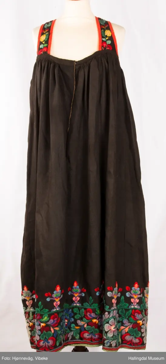Frihåndsbrodert festbunad fra Nedre Hallingdal. Stakken er sydd av svart ulltøy, toskaftvevd av 1-tråds garn (grannty). 5 like brede høyder i stakken som er rynket øverst så nær som et ørlite stykke midt framme. På stakken er festet ei list øverst. Fyldig hallingsaum-broderi, akantusranke med 11 roser som er brodert rett på stakken. Over akantusranken er det broderte greiner som står loddrett på en brodert grønn rett bord. Nederst på stakken er det en slitekant av grønt ullbånd som er brettet inn på baksiden. Over denne er det røde kroklisser. Stakken er skonet med et gråbrunt kypertvevd bomullstoff.  Overlivet er foret med hvitt bomullslerret, en ørliten stikklomme (h. 7 cm, br. 4,7 cm) på innsiden av det ene framstykket. Lomma er av rutete gult, brunt og blått bomullstøy. Overlivet er kantet med røde ullbånd. Det går sammen under ermene med sidesømmer på 4,5 cm her, bare 0,6 cm ovom lista (resten er nedom lista på vranga). Stakketøyet er håndvevd, trolig også foret i overlivet. Stoff og bånd er ellers fabrikkvevd.  Brodert for hånd. Høydene i stakken er sydd sammen for hånd.  Noe maskinsøm på kantebåndene.