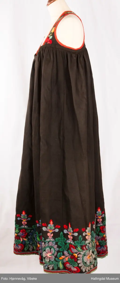 Frihåndsbrodert festbunad fra Nedre Hallingdal. Stakken er sydd av svart ulltøy, toskaftvevd av 1-tråds garn (grannty). 5 like brede høyder i stakken som er rynket øverst så nær som et ørlite stykke midt framme. På stakken er festet ei list øverst. Fyldig hallingsaum-broderi, akantusranke med 11 roser som er brodert rett på stakken. Over akantusranken er det broderte greiner som står loddrett på en brodert grønn rett bord. Nederst på stakken er det en slitekant av grønt ullbånd som er brettet inn på baksiden. Over denne er det røde kroklisser. Stakken er skonet med et gråbrunt kypertvevd bomullstoff.  Overlivet er foret med hvitt bomullslerret, en ørliten stikklomme (h. 7 cm, br. 4,7 cm) på innsiden av det ene framstykket. Lomma er av rutete gult, brunt og blått bomullstøy. Overlivet er kantet med røde ullbånd. Det går sammen under ermene med sidesømmer på 4,5 cm her, bare 0,6 cm ovom lista (resten er nedom lista på vranga). Stakketøyet er håndvevd, trolig også foret i overlivet. Stoff og bånd er ellers fabrikkvevd.  Brodert for hånd. Høydene i stakken er sydd sammen for hånd.  Noe maskinsøm på kantebåndene.