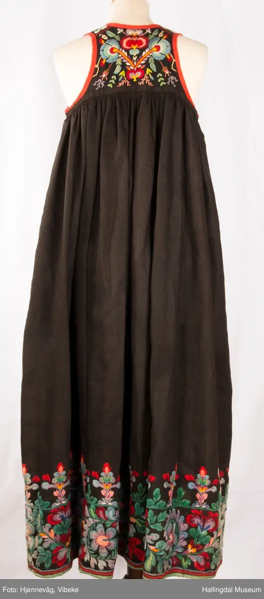 Frihåndsbrodert festbunad fra Nedre Hallingdal. Stakken er sydd av svart ulltøy, toskaftvevd av 1-tråds garn (grannty). 5 like brede høyder i stakken som er rynket øverst så nær som et ørlite stykke midt framme. På stakken er festet ei list øverst. Fyldig hallingsaum-broderi, akantusranke med 11 roser som er brodert rett på stakken. Over akantusranken er det broderte greiner som står loddrett på en brodert grønn rett bord. Nederst på stakken er det en slitekant av grønt ullbånd som er brettet inn på baksiden. Over denne er det røde kroklisser. Stakken er skonet med et gråbrunt kypertvevd bomullstoff.  Overlivet er foret med hvitt bomullslerret, en ørliten stikklomme (h. 7 cm, br. 4,7 cm) på innsiden av det ene framstykket. Lomma er av rutete gult, brunt og blått bomullstøy. Overlivet er kantet med røde ullbånd. Det går sammen under ermene med sidesømmer på 4,5 cm her, bare 0,6 cm ovom lista (resten er nedom lista på vranga). Stakketøyet er håndvevd, trolig også foret i overlivet. Stoff og bånd er ellers fabrikkvevd.  Brodert for hånd. Høydene i stakken er sydd sammen for hånd.  Noe maskinsøm på kantebåndene.