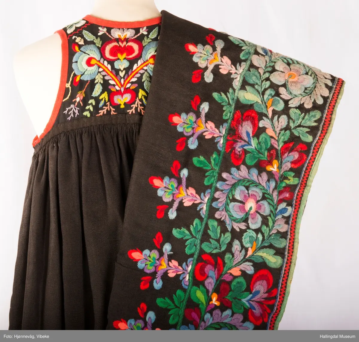 Frihåndsbrodert festbunad fra Nedre Hallingdal. Stakken er sydd av svart ulltøy, toskaftvevd av 1-tråds garn (grannty). 5 like brede høyder i stakken som er rynket øverst så nær som et ørlite stykke midt framme. På stakken er festet ei list øverst. Fyldig hallingsaum-broderi, akantusranke med 11 roser som er brodert rett på stakken. Over akantusranken er det broderte greiner som står loddrett på en brodert grønn rett bord. Nederst på stakken er det en slitekant av grønt ullbånd som er brettet inn på baksiden. Over denne er det røde kroklisser. Stakken er skonet med et gråbrunt kypertvevd bomullstoff.  Overlivet er foret med hvitt bomullslerret, en ørliten stikklomme (h. 7 cm, br. 4,7 cm) på innsiden av det ene framstykket. Lomma er av rutete gult, brunt og blått bomullstøy. Overlivet er kantet med røde ullbånd. Det går sammen under ermene med sidesømmer på 4,5 cm her, bare 0,6 cm ovom lista (resten er nedom lista på vranga). Stakketøyet er håndvevd, trolig også foret i overlivet. Stoff og bånd er ellers fabrikkvevd.  Brodert for hånd. Høydene i stakken er sydd sammen for hånd.  Noe maskinsøm på kantebåndene.