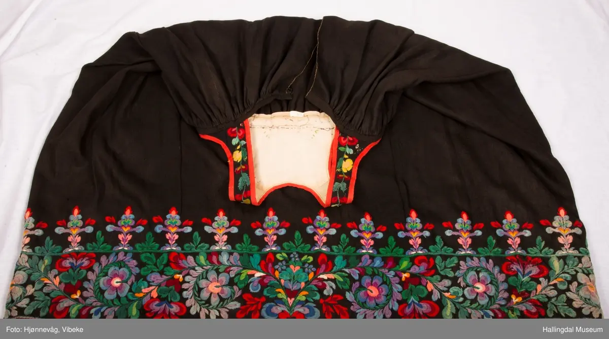 Frihåndsbrodert festbunad fra Nedre Hallingdal. Stakken er sydd av svart ulltøy, toskaftvevd av 1-tråds garn (grannty). 5 like brede høyder i stakken som er rynket øverst så nær som et ørlite stykke midt framme. På stakken er festet ei list øverst. Fyldig hallingsaum-broderi, akantusranke med 11 roser som er brodert rett på stakken. Over akantusranken er det broderte greiner som står loddrett på en brodert grønn rett bord. Nederst på stakken er det en slitekant av grønt ullbånd som er brettet inn på baksiden. Over denne er det røde kroklisser. Stakken er skonet med et gråbrunt kypertvevd bomullstoff.  Overlivet er foret med hvitt bomullslerret, en ørliten stikklomme (h. 7 cm, br. 4,7 cm) på innsiden av det ene framstykket. Lomma er av rutete gult, brunt og blått bomullstøy. Overlivet er kantet med røde ullbånd. Det går sammen under ermene med sidesømmer på 4,5 cm her, bare 0,6 cm ovom lista (resten er nedom lista på vranga). Stakketøyet er håndvevd, trolig også foret i overlivet. Stoff og bånd er ellers fabrikkvevd.  Brodert for hånd. Høydene i stakken er sydd sammen for hånd.  Noe maskinsøm på kantebåndene.