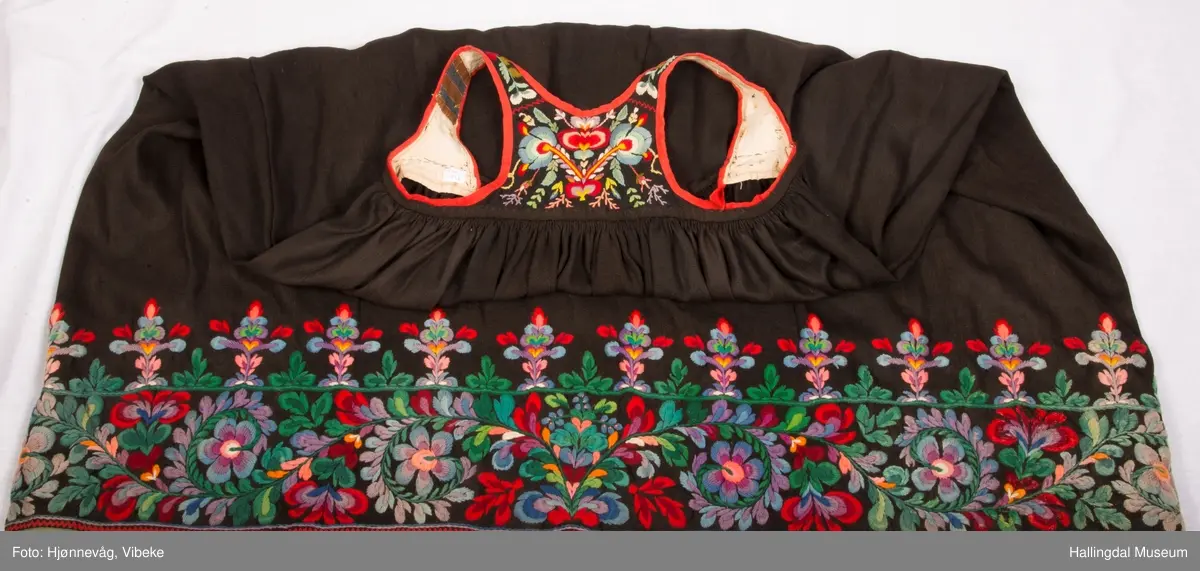 Frihåndsbrodert festbunad fra Nedre Hallingdal. Stakken er sydd av svart ulltøy, toskaftvevd av 1-tråds garn (grannty). 5 like brede høyder i stakken som er rynket øverst så nær som et ørlite stykke midt framme. På stakken er festet ei list øverst. Fyldig hallingsaum-broderi, akantusranke med 11 roser som er brodert rett på stakken. Over akantusranken er det broderte greiner som står loddrett på en brodert grønn rett bord. Nederst på stakken er det en slitekant av grønt ullbånd som er brettet inn på baksiden. Over denne er det røde kroklisser. Stakken er skonet med et gråbrunt kypertvevd bomullstoff.  Overlivet er foret med hvitt bomullslerret, en ørliten stikklomme (h. 7 cm, br. 4,7 cm) på innsiden av det ene framstykket. Lomma er av rutete gult, brunt og blått bomullstøy. Overlivet er kantet med røde ullbånd. Det går sammen under ermene med sidesømmer på 4,5 cm her, bare 0,6 cm ovom lista (resten er nedom lista på vranga). Stakketøyet er håndvevd, trolig også foret i overlivet. Stoff og bånd er ellers fabrikkvevd.  Brodert for hånd. Høydene i stakken er sydd sammen for hånd.  Noe maskinsøm på kantebåndene.
