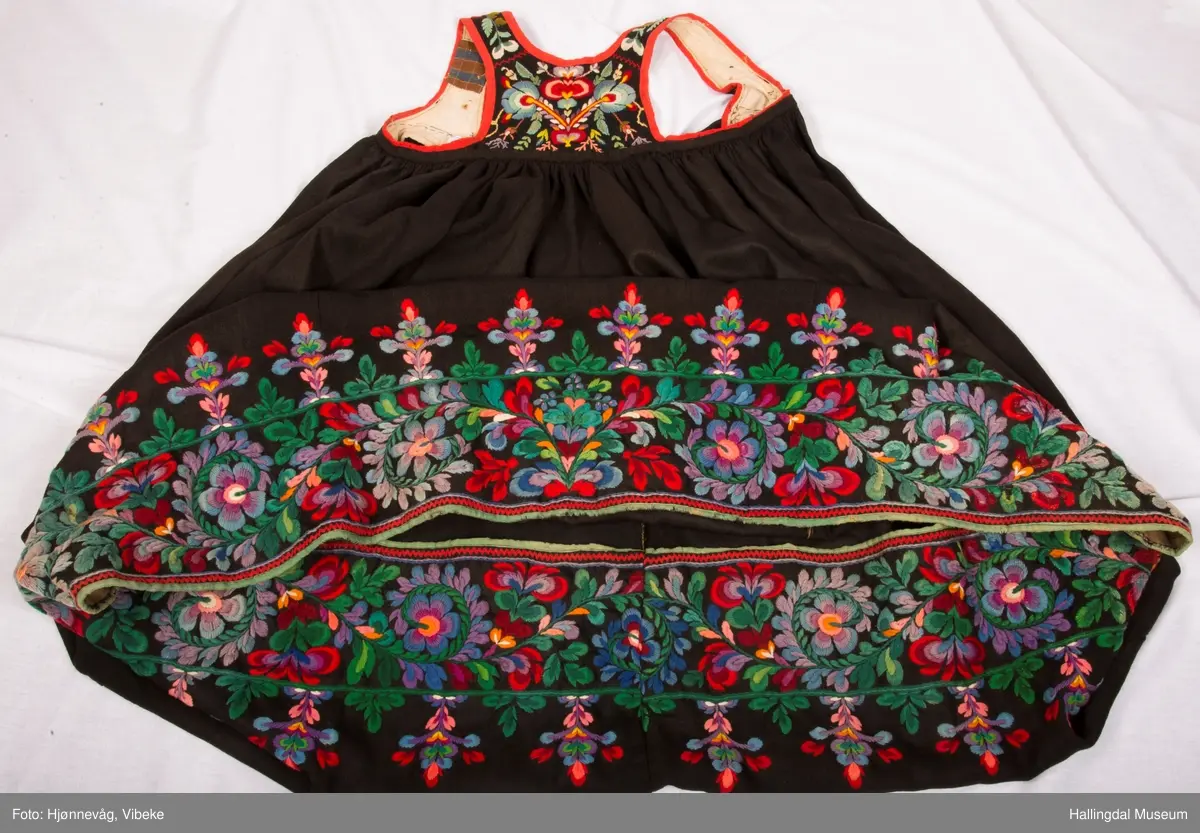Frihåndsbrodert festbunad fra Nedre Hallingdal. Stakken er sydd av svart ulltøy, toskaftvevd av 1-tråds garn (grannty). 5 like brede høyder i stakken som er rynket øverst så nær som et ørlite stykke midt framme. På stakken er festet ei list øverst. Fyldig hallingsaum-broderi, akantusranke med 11 roser som er brodert rett på stakken. Over akantusranken er det broderte greiner som står loddrett på en brodert grønn rett bord. Nederst på stakken er det en slitekant av grønt ullbånd som er brettet inn på baksiden. Over denne er det røde kroklisser. Stakken er skonet med et gråbrunt kypertvevd bomullstoff.  Overlivet er foret med hvitt bomullslerret, en ørliten stikklomme (h. 7 cm, br. 4,7 cm) på innsiden av det ene framstykket. Lomma er av rutete gult, brunt og blått bomullstøy. Overlivet er kantet med røde ullbånd. Det går sammen under ermene med sidesømmer på 4,5 cm her, bare 0,6 cm ovom lista (resten er nedom lista på vranga). Stakketøyet er håndvevd, trolig også foret i overlivet. Stoff og bånd er ellers fabrikkvevd.  Brodert for hånd. Høydene i stakken er sydd sammen for hånd.  Noe maskinsøm på kantebåndene.