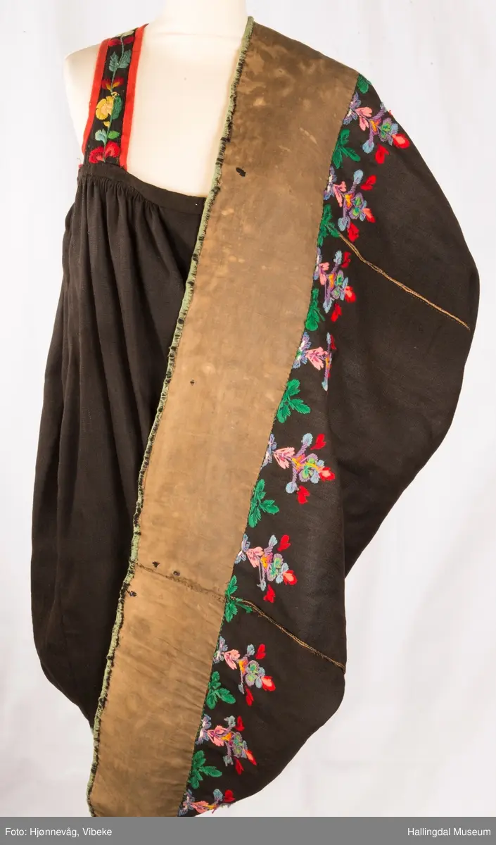 Frihåndsbrodert festbunad fra Nedre Hallingdal. Stakken er sydd av svart ulltøy, toskaftvevd av 1-tråds garn (grannty). 5 like brede høyder i stakken som er rynket øverst så nær som et ørlite stykke midt framme. På stakken er festet ei list øverst. Fyldig hallingsaum-broderi, akantusranke med 11 roser som er brodert rett på stakken. Over akantusranken er det broderte greiner som står loddrett på en brodert grønn rett bord. Nederst på stakken er det en slitekant av grønt ullbånd som er brettet inn på baksiden. Over denne er det røde kroklisser. Stakken er skonet med et gråbrunt kypertvevd bomullstoff.  Overlivet er foret med hvitt bomullslerret, en ørliten stikklomme (h. 7 cm, br. 4,7 cm) på innsiden av det ene framstykket. Lomma er av rutete gult, brunt og blått bomullstøy. Overlivet er kantet med røde ullbånd. Det går sammen under ermene med sidesømmer på 4,5 cm her, bare 0,6 cm ovom lista (resten er nedom lista på vranga). Stakketøyet er håndvevd, trolig også foret i overlivet. Stoff og bånd er ellers fabrikkvevd.  Brodert for hånd. Høydene i stakken er sydd sammen for hånd.  Noe maskinsøm på kantebåndene.