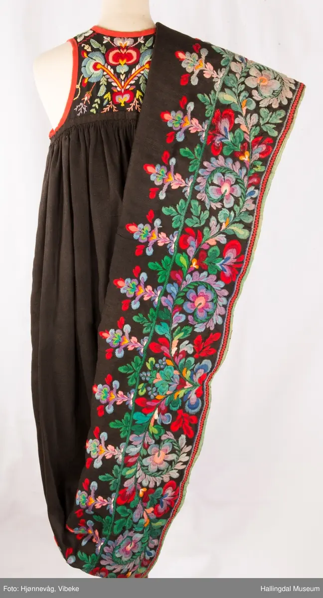 Frihåndsbrodert festbunad fra Nedre Hallingdal. Stakken er sydd av svart ulltøy, toskaftvevd av 1-tråds garn (grannty). 5 like brede høyder i stakken som er rynket øverst så nær som et ørlite stykke midt framme. På stakken er festet ei list øverst. Fyldig hallingsaum-broderi, akantusranke med 11 roser som er brodert rett på stakken. Over akantusranken er det broderte greiner som står loddrett på en brodert grønn rett bord. Nederst på stakken er det en slitekant av grønt ullbånd som er brettet inn på baksiden. Over denne er det røde kroklisser. Stakken er skonet med et gråbrunt kypertvevd bomullstoff.  Overlivet er foret med hvitt bomullslerret, en ørliten stikklomme (h. 7 cm, br. 4,7 cm) på innsiden av det ene framstykket. Lomma er av rutete gult, brunt og blått bomullstøy. Overlivet er kantet med røde ullbånd. Det går sammen under ermene med sidesømmer på 4,5 cm her, bare 0,6 cm ovom lista (resten er nedom lista på vranga). Stakketøyet er håndvevd, trolig også foret i overlivet. Stoff og bånd er ellers fabrikkvevd.  Brodert for hånd. Høydene i stakken er sydd sammen for hånd.  Noe maskinsøm på kantebåndene.