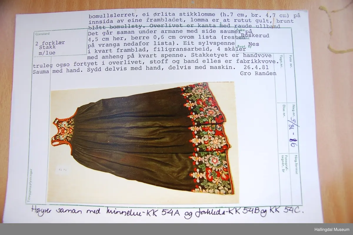 Frihåndsbrodert festbunad fra Nedre Hallingdal. Stakken er sydd av svart ulltøy, toskaftvevd av 1-tråds garn (grannty). 5 like brede høyder i stakken som er rynket øverst så nær som et ørlite stykke midt framme. På stakken er festet ei list øverst. Fyldig hallingsaum-broderi, akantusranke med 11 roser som er brodert rett på stakken. Over akantusranken er det broderte greiner som står loddrett på en brodert grønn rett bord. Nederst på stakken er det en slitekant av grønt ullbånd som er brettet inn på baksiden. Over denne er det røde kroklisser. Stakken er skonet med et gråbrunt kypertvevd bomullstoff.  Overlivet er foret med hvitt bomullslerret, en ørliten stikklomme (h. 7 cm, br. 4,7 cm) på innsiden av det ene framstykket. Lomma er av rutete gult, brunt og blått bomullstøy. Overlivet er kantet med røde ullbånd. Det går sammen under ermene med sidesømmer på 4,5 cm her, bare 0,6 cm ovom lista (resten er nedom lista på vranga). Stakketøyet er håndvevd, trolig også foret i overlivet. Stoff og bånd er ellers fabrikkvevd.  Brodert for hånd. Høydene i stakken er sydd sammen for hånd.  Noe maskinsøm på kantebåndene.