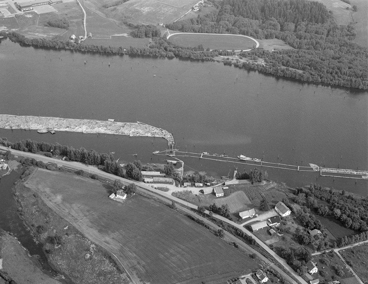 Flyfotografi som viser en del av lenseanlegget ved Fetsund i Akershus, der løstømmer som kom flytende fra den øvre delen av Glommavassdraget ble samlet og «soppet» (buntet) med sikte på buksering videre sørover innsjøen Øyeren med en slepebåt som trekkraft. Dette bildet er tatt fra en posisjon over den vestre elvebredden, med Glomma fellesfløtingsforenings eiendom Lund i forgrunnen. Ute på vannspeilet ser vi det såkalte retteanlegget, der tømmer fra ei traktformet lense som var krum i den nedre enden ble trukket over ei bru med piggvalser og kastet parallelt ned i tømmerrenna. Sentralt i denne renna gikk det en vaier som trakk det langsomt nedover mot «mosemaskinen» nederst i anlegget, der det ble buntet. Mosemaskinen er ikke med på dette fotografiet. Derimot lå en del av båtene som ble brukt ved lenseanlegget fortøyd på yttersida av tømmerrenna. Bildet ble tatt i 1984, den nest siste sesongen det var fløting i Glomma. Da var tømmervolumet såpass lite at det bare var en mosemaskin som fortsatt var i drift. På dette tidspunktet ble anlegget drevet av et mannskap på cirka 30. Mer informasjon om lensedriften ved Fetsund finnes under fanen «Opplysninger».