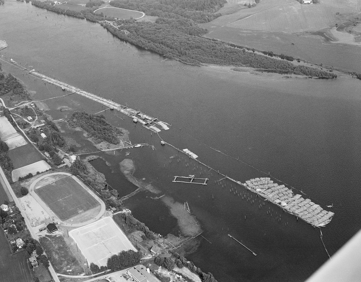 Flyfotografi som viser en del av lenseanlegget ved Fetsund i Akershus, der løstømmer som kom flytende fra den øvre delen av Glommavassdraget ble samlet og «soppet» (buntet) med sikte på buksering videre sørover innsjøen Øyeren med en slepebåt som trekkraft. Dette bildet er tatt fra en posisjon over Øya, landtangen som skilte Glommas utløp i Øyeren fra Svellet, en innsjø som hadde tilløp fra Nitelva. På Glommas vannspeil ser vi den nedre delen av den såkalte Lundslensa ved vestre elvebredd.  Her ble løstømmeret fra den ovenforliggende delen av vassdraget ledet via et retteanlegg inn i ei tømmerrenne, en åpen kanal omgitt av flåteganger på begge sider. Den nedre delen av denne renna var todelt - tømmeret kunne styres mot den ene eller den andre delen ved hjelp av en manuell pens. I ytterendene av de to rennene var det «soppemaskiner» som presset tømmeret sammen i «sopper», bunter, som etter hvert skulle slepes sørover innsjøen Øyeren. I deltaområdet like nedenfor lenseanlegget var det grunt, og på denne strekningen måtte slepebåtene manøvreres forsiktig over djupålene på sand- og slambankene. I dette området kunne slepene bestå av cirka 200 «sopper». Da dette fotografiet ble tatt lå et slikt slep på vent i et innlenset område like nedenfor lenseanlegget. Til venstre i forgrunnen ser vi stadionet til det lokale idrettslaget. Mer informasjon om lensedriften ved Fetsund finnes under fanen «Opplysninger».