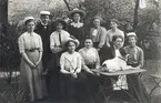 En grupp kvinnor i studentmössor m.m. tillsammans med en man och en hund, avfotograferade i 
en trädgård i Lund (?), 1912.