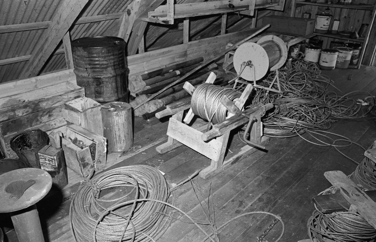 Fra loftet over båthuset ved Sørlistøa fløtermuseum ved Osensjøen i Åmot i Hedmark. Dette er en bortimot 25 meter lang og drøyt 8 meter bred bygning, oppført i bordkledd bindingsverk, og med bølgeblikktekket saltak. Båtene ble tatt inn og ut av huset av bygningen via porter i den nordvestre bygningsgavlen, dit det gikk en skinnegang på bakkeplan fra slippen. Dette fotografiet er tatt fra loftet over denne nordvestre delen av bygningen, der Glomma fellesfløtingsforenings mannskaper lagret diverse materiell de trengte under arbeidet med å buksere tømmer over Osensjøen. Slepetrosse, Wirevinde, Tjæretønne. Fra loftet på Sørlistøa ved Osensjøen, Nordre Osen, Åmot, Hedmark.