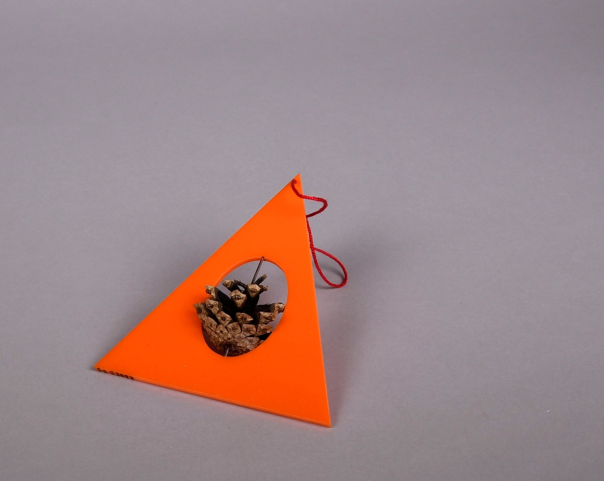 Juletrefigur av farget akryl og furukongle. Juletre formet som oransjefarget trekant med ovalt hull i midten, i hullet er det en stav av metall som går gjennom en furukongle.
